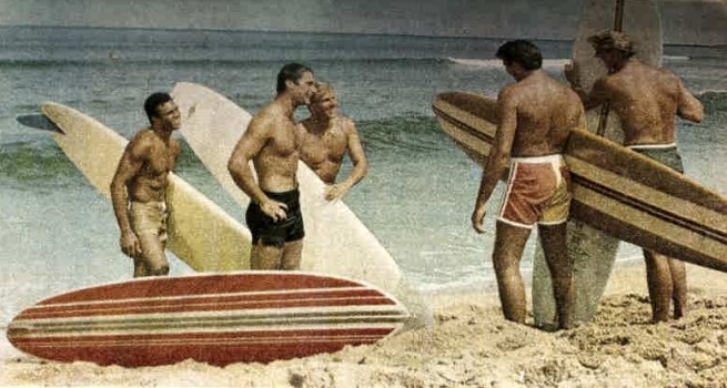 The image is a still from the 1964 film Ride the Wild Surf and shows four white men surfers and one man of colour standing on the shore of the beach. They are holding surfboards. The four white men are all looking at one another, the man of colour is on the outside of the group.