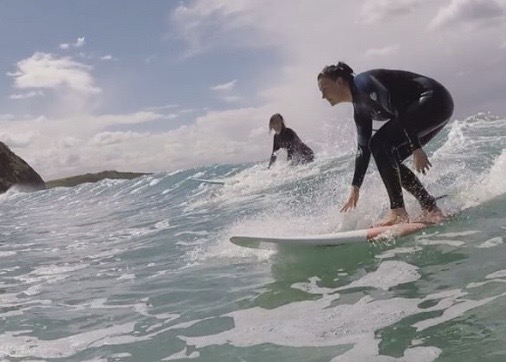 Daniella popping up on a wave with a fellow surfer in the background.