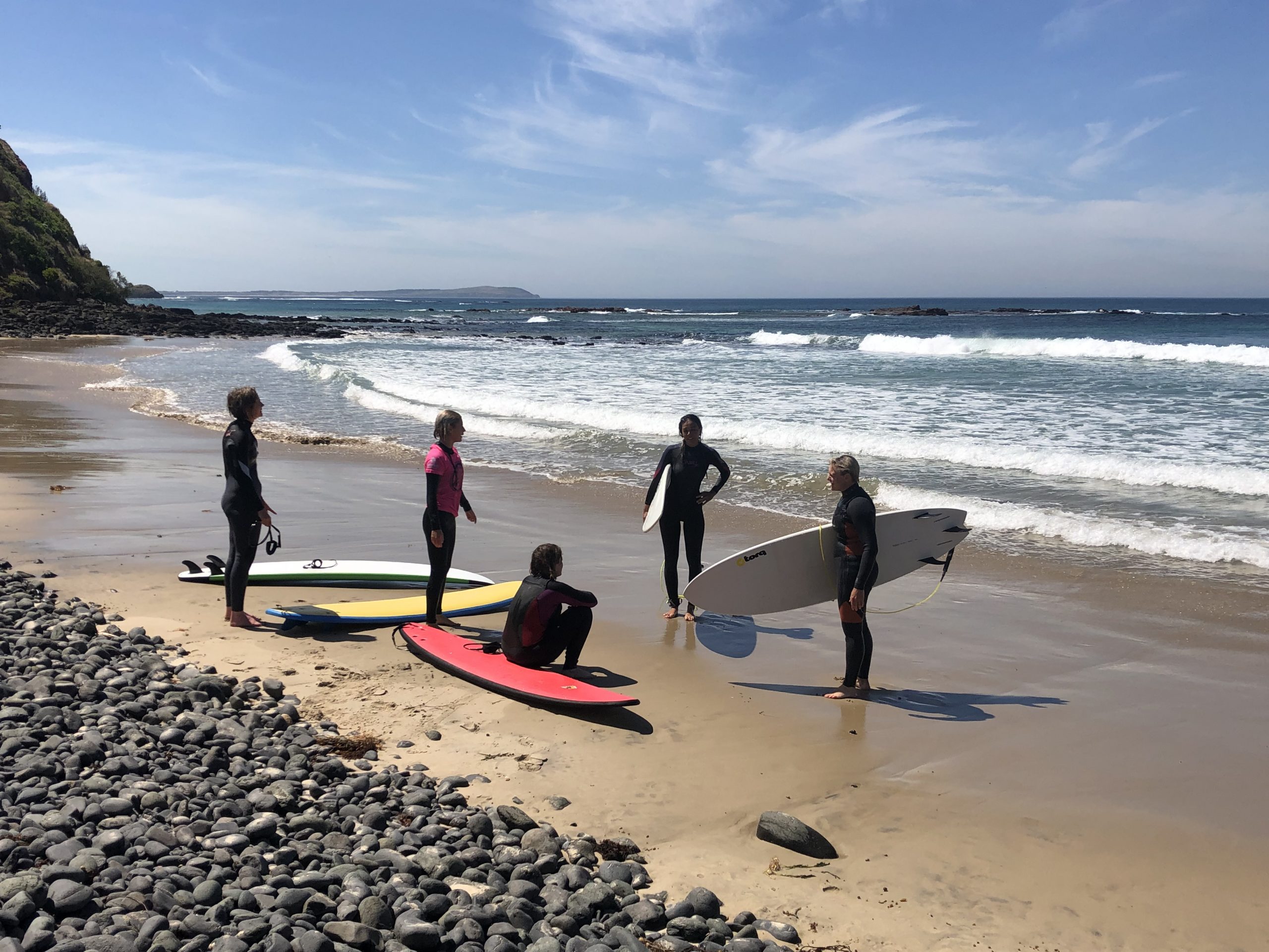 Jess and four of her surf students standing on the shore with their surfboards after a surf. The weather is sunny.