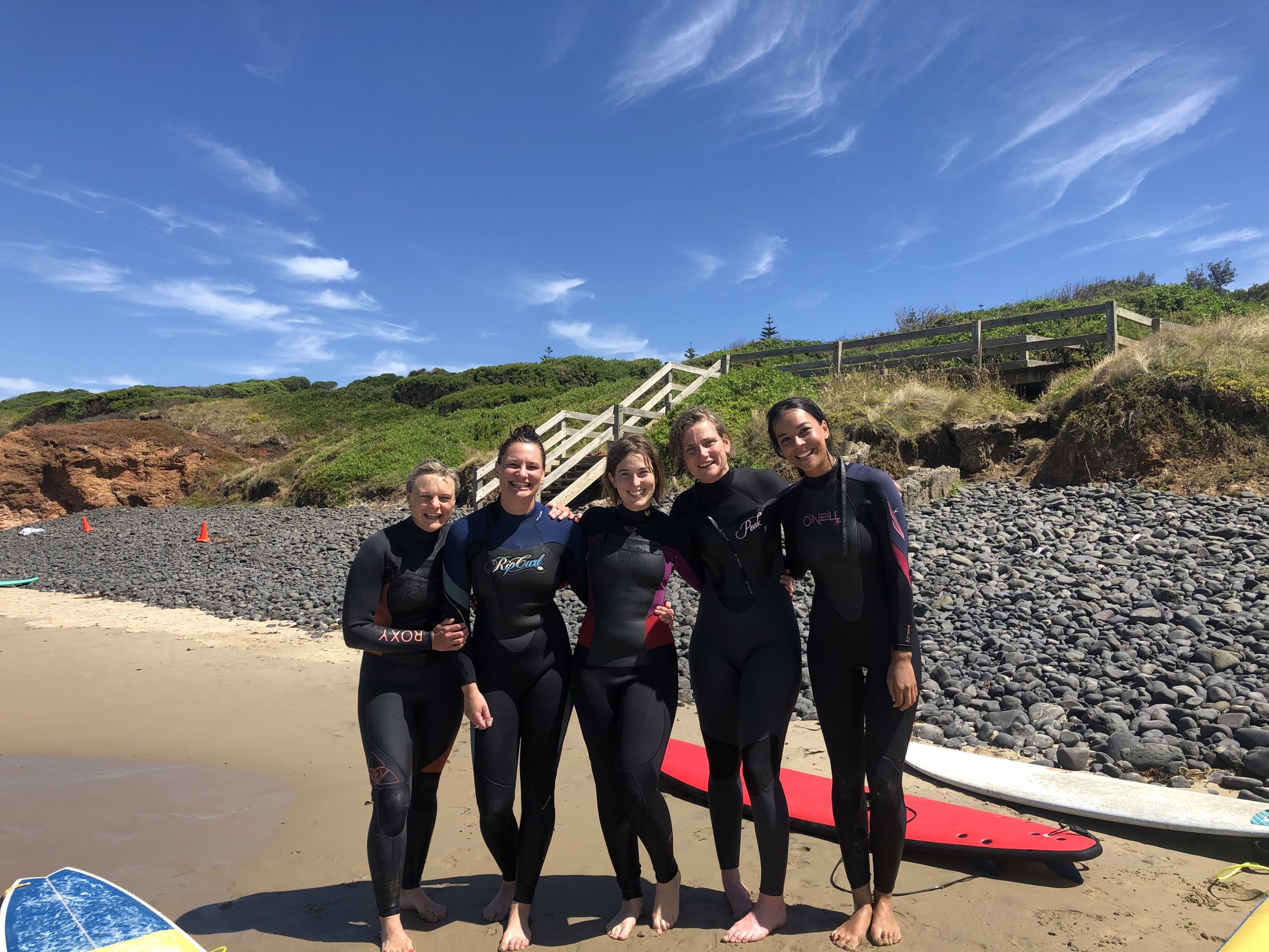 Five surf school students standing on the shore after a surf session. They are all smiling.