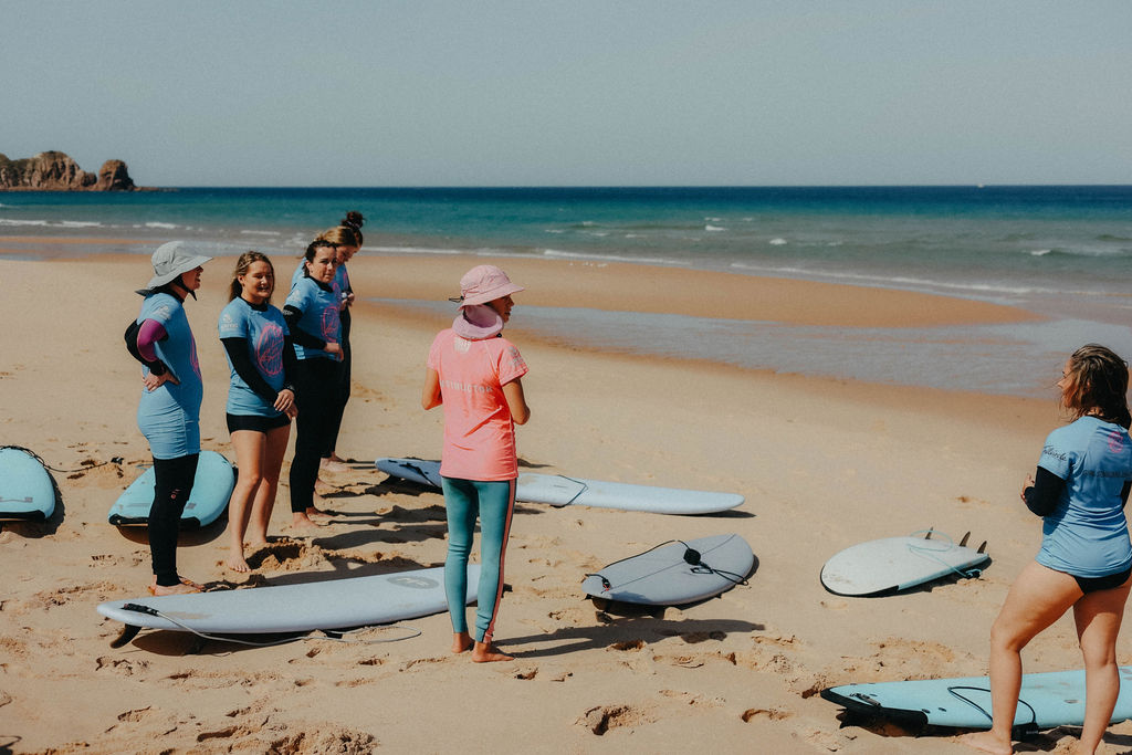 Jess in coach mode: standing on the beach talking to six students. Jess is wearing a blue wetsuit with pink rash vest; the students are wearing blue Girls on Board rash vests. Their surfboards are on the sand in preparation for a surf.