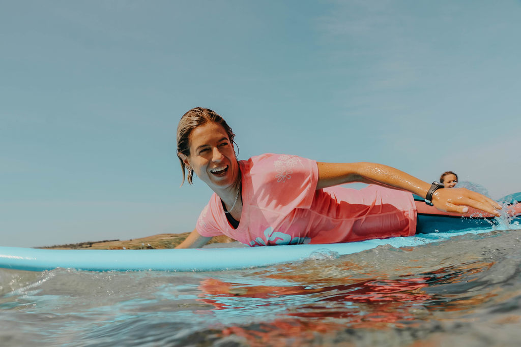 Jess paddling on a blue board wearing a pastel pink rash vest. She is smiling and the sky is clear blue. 