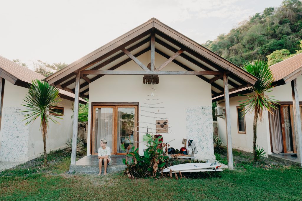 Jess sitting on the pergola floor of a bungalow in a tropical location with palm trees either side. She is wearing a beige t-shirt and looking to the side of the frame. Two surfboards are stacked on top of each other next to her and some ferns.