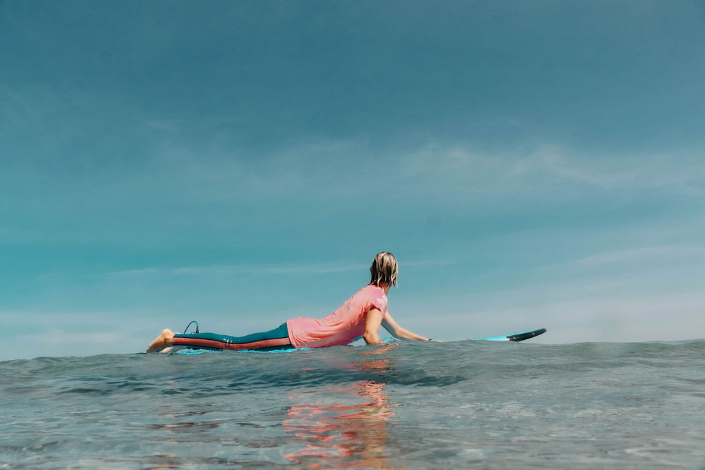 Jess paddling on her surfboard in the ocean. She is wearing a blue wetsuit and a pink rash vest. The ocean is calm and the sky blue. Jess is looking away from the camera.