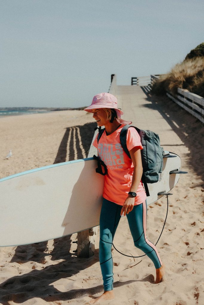 Jess walking onto the beach on a sunny day, holding a surfboard. She is wearing a blue wetsuit, a pink Girls on Board rash vest and a pink cap. She is smiling.