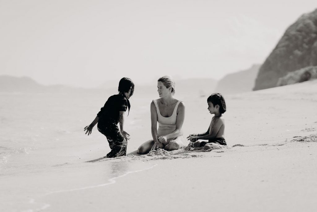 A black and white photo of Jess sitting on the shore playing in the sand with two young children. Jess is wearing a one piece swimsuit and her hair is slicked back.