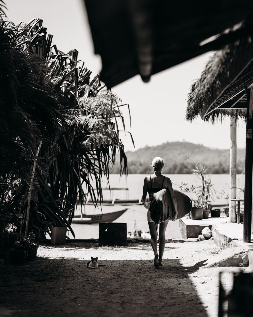 A black and white image of Jess walking towards the ocean in a tropical location. She is wearing shorts and a singlet, carrying a surfboard under one arm, and walking away from the camera. A small black and white cat sits on the ground facing the camera.