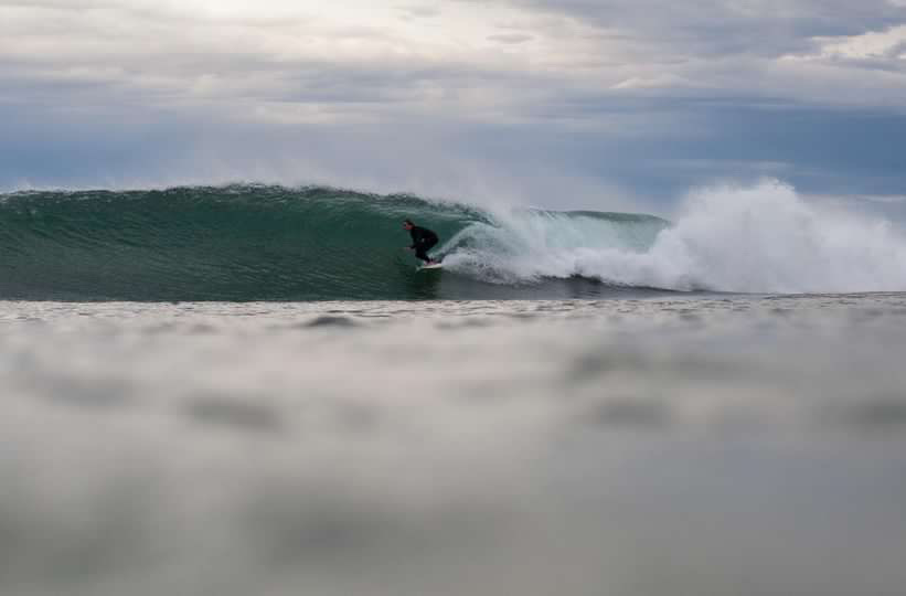 Image of Linley getting barrelled in a right hander wave. She is wearing a full steamer wetsuit and the sky above her and the wave is grey and cloudy.