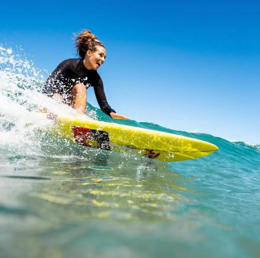 Linley surfing a left hander, smiling. She is wearing a black springsuit and riding a yellow surfboard with a sticker of the Aboriginal flag on the bottom. The sky is clear and blue.