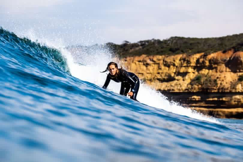 Image of Linley surfing a right hander wave. She is looking down the line towards the photographer. There are cliffs in the background. Linely is wearing a full steamer wetsuit.