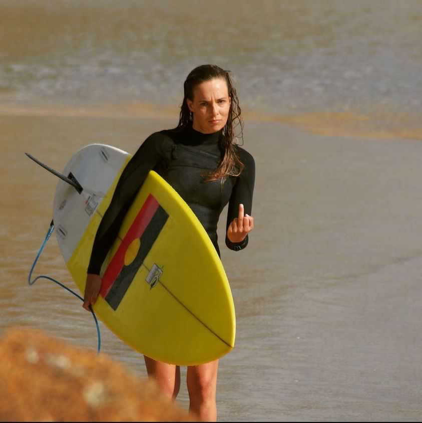 Linley walking along the beach holding a yellow surfboard with a sticker of the Aboriginal flag on the bottom. She is giving the finger in the direction of the camera.