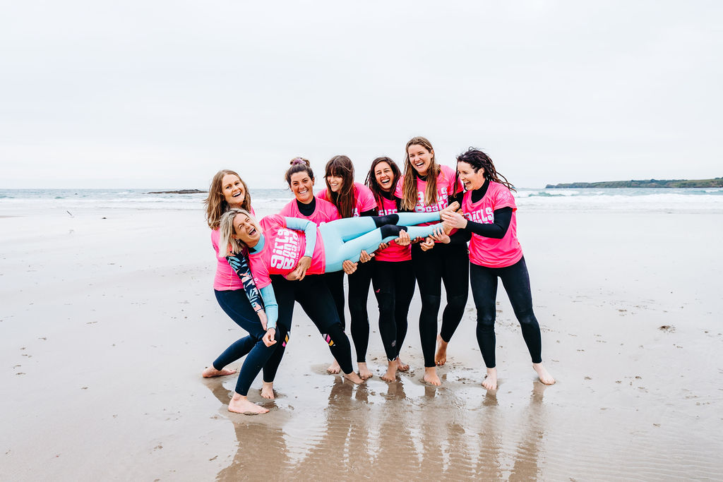 Jess being held off the ground, sideways, by her team of women surf coaches. Jess is wearing a blue wetsuit and those holding her are wearing black wetsuits. They are all laughing and wearing pink Girls on Board rash vests. They are standing on the shore with the ocean behind them.